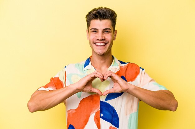 Молодой кавказский мужчина на желтом фоне улыбается и показывает руками форму сердца.