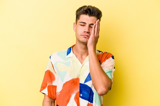 Young caucasian man isolated on yellow background tired and very sleepy keeping hand on head