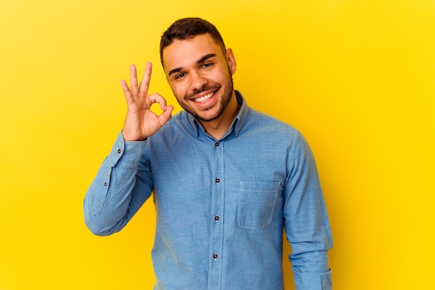 Young caucasian man isolated on yellow background winks an eye and holds an okay gesture with hand.