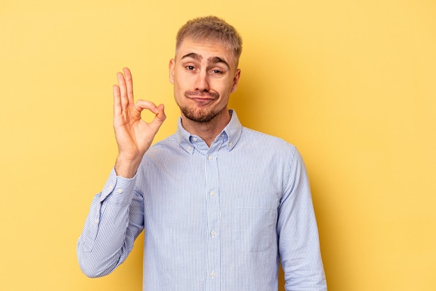 Young caucasian man isolated on yellow background winks an eye and holds an okay gesture with hand.