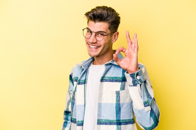 Young caucasian man isolated on yellow background winks an eye and holds an okay gesture with hand.