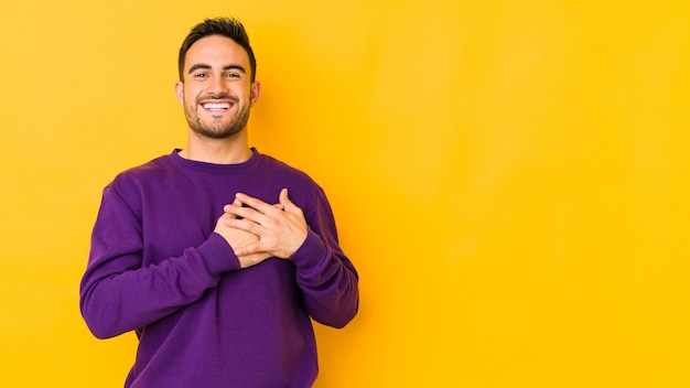 Young caucasian man isolated on yellow bakground laughing keeping hands on heart, concept of happiness.