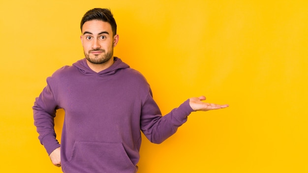 Young caucasian man isolated on yellow bakground showing a copy space on a palm and holding another hand on waist.