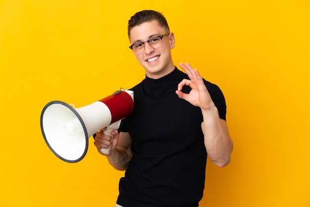 Young caucasian man isolated on yellow holding a megaphone and showing ok sign with fingers