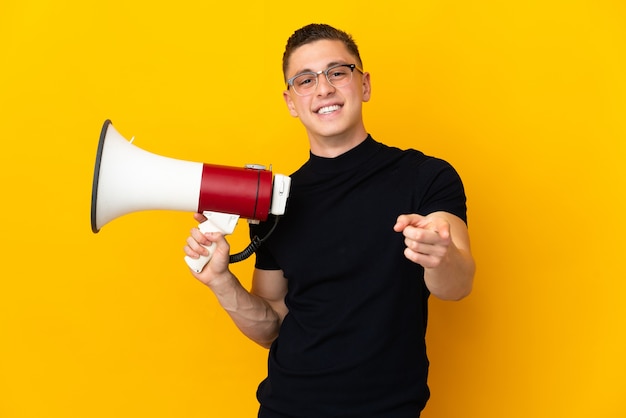 Young caucasian man isolated on yellow holding a megaphone and smiling while pointing to the front