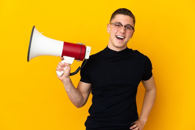 Young caucasian man isolated on yellow holding a megaphone and smiling