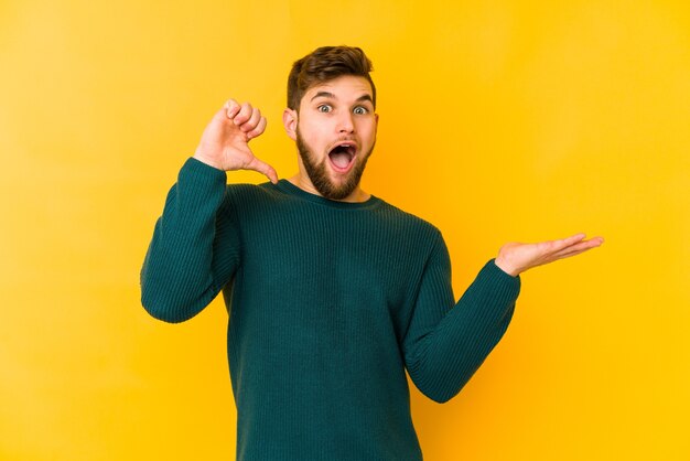 Young caucasian man isolated on yellow holds copy space on palm, keep hand on cheek. Amazed and delighted.