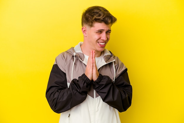 Young caucasian man isolated on yellow wall holding hands in pray near mouth, feels confident.