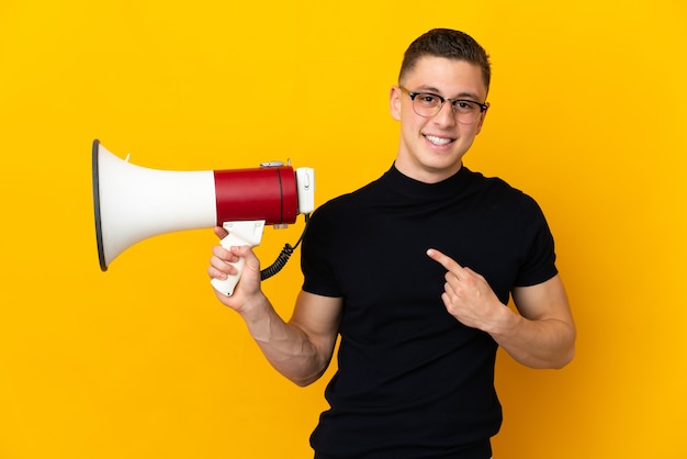 Young caucasian man isolated on yellow wall holding a megaphone and pointing side