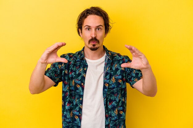 Young caucasian man isolated on yellow wall holding something with palms