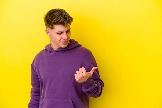 Young caucasian man isolated on yellow wall points with thumb finger away, laughing and carefree