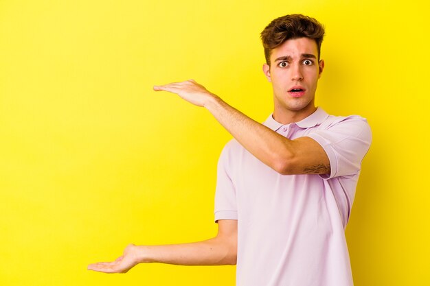 Young caucasian man isolated on yellow wall shocked and amazed holding a copy space between hands