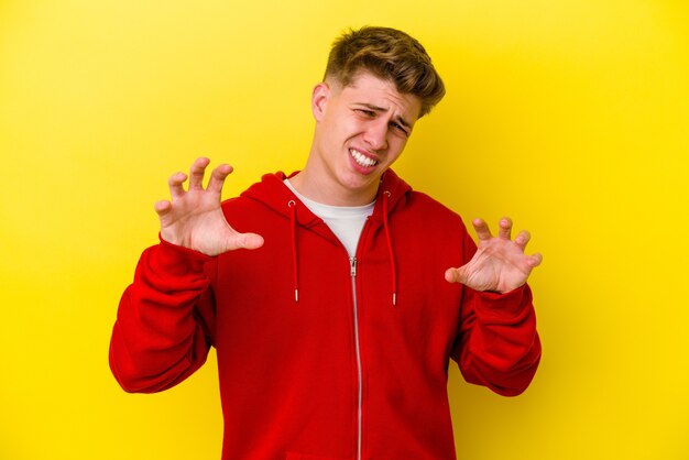 Young caucasian man isolated on yellow wall showing claws imitating a cat, aggressive gesture.