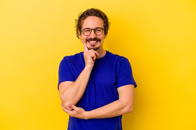 Young caucasian man isolated on yellow wall smiling happy and confident, touching chin with hand