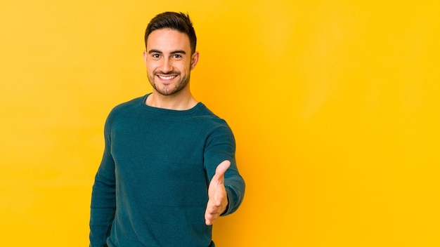 Giovane uomo caucasico isolato sulla parete gialla che allunga la mano nel gesto di saluto.