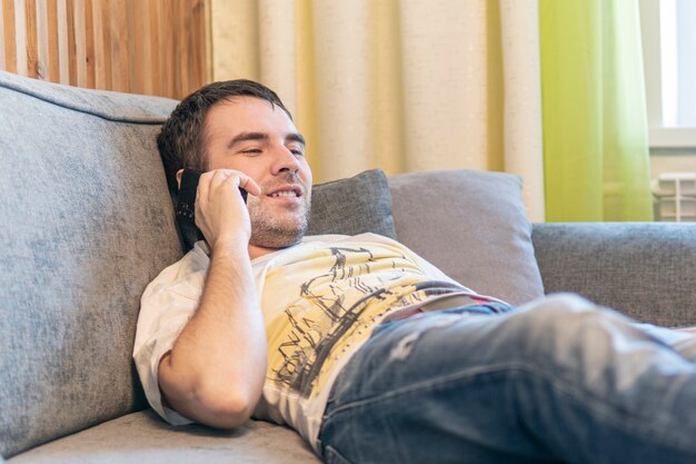 若い白人男性が自宅のソファでリラックスして横たわる現代のスマートフォンガジェット、リビングルームの快適なソファでリラックスしたミレニアル世代の男性の休息、技術コンセプト