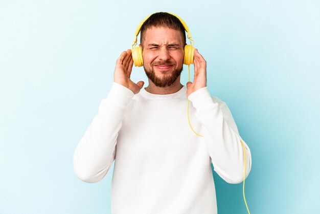 Young caucasian man listening to music isolated on blue background covering ears with hands.