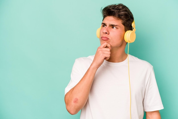 Young caucasian man listening to music isolated on blue background looking sideways with doubtful and skeptical expression