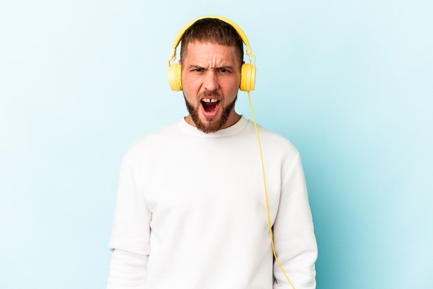 Young caucasian man listening to music isolated on blue background screaming very angry and aggressive.