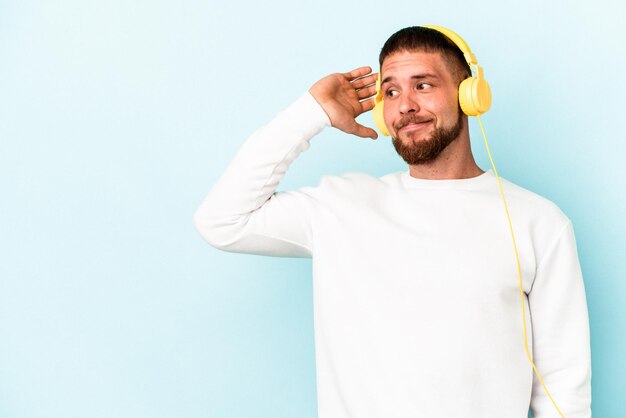 Giovane uomo caucasico che ascolta musica isolato su sfondo blu toccando la parte posteriore della testa, pensando e facendo una scelta.