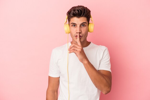 Young caucasian man listening to music isolated on pink background keeping a secret or asking for silence.