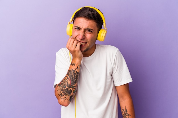 Young caucasian man listening to music isolated on purple background  biting fingernails, nervous and very anxious.