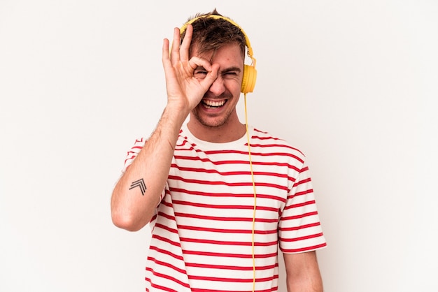 Young caucasian man listening to music isolated on white background excited keeping ok gesture on eye.