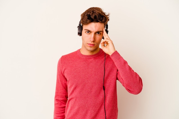 Young caucasian man listening to music isolated on white background pointing temple with finger, thinking, focused on a task.