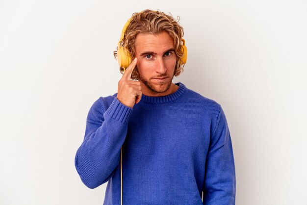 Young caucasian man listening to music isolated on white background pointing temple with finger, thinking, focused on a task.