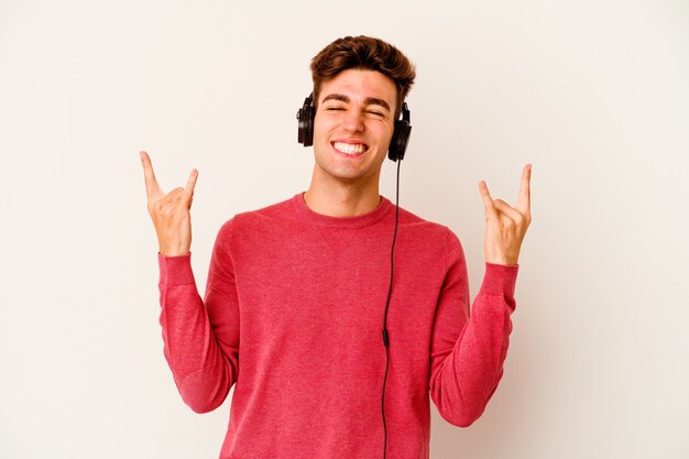 Giovane uomo caucasico che ascolta musica isolato su sfondo bianco che mostra gesto rock con le dita