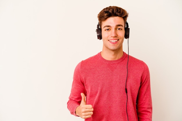 Giovane uomo caucasico ascoltando musica isolato su sfondo bianco sorridendo e alzando il pollice