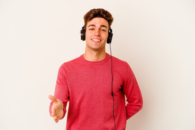 Young caucasian man listening to music isolated on white background stretching hand at camera in greeting gesture.