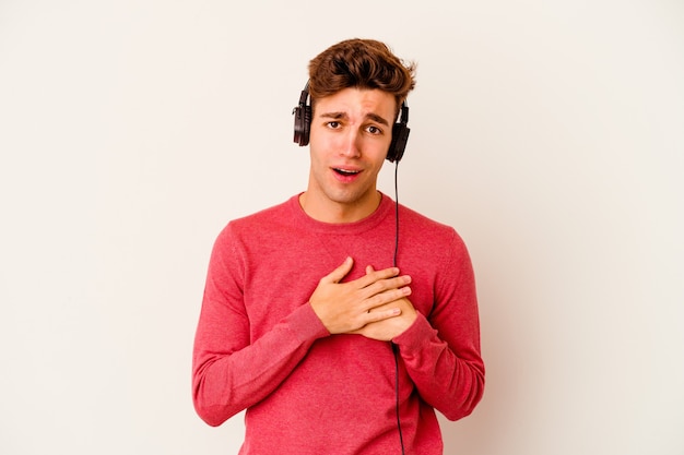 Young caucasian man listening to music isolated on white wall has friendly expression, pressing palm to chest. Love concept.