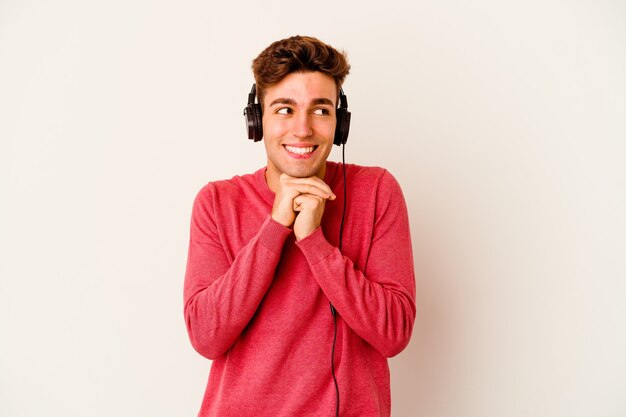 Young caucasian man listening to music isolated on white wall keeps hands under chin, is looking happily aside.