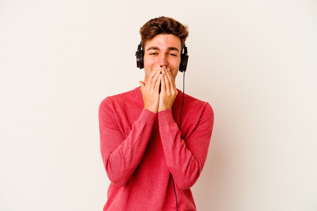 Young caucasian man listening to music isolated on white wall laughing about something, covering mouth with hands