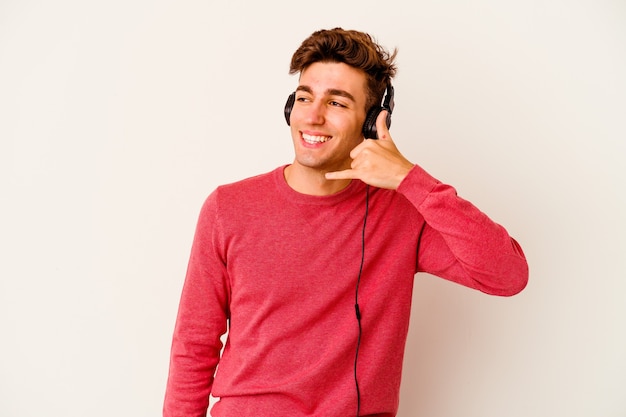 Young caucasian man listening to music isolated on white wall showing a mobile phone call gesture with fingers.