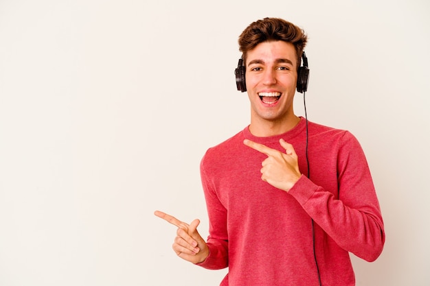 Young caucasian man listening to music on white pointing with forefingers to a copy space, expressing excitement and desire.