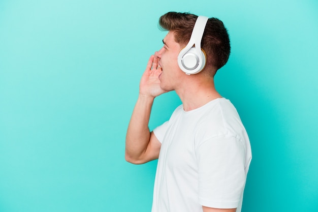 Young caucasian man listening to music with headphones isolated on blue wall shouting and holding palm near opened mouth