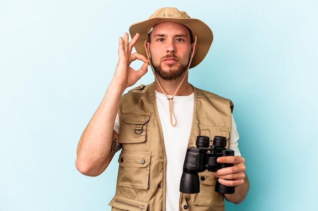 Young caucasian man looking at animals through binoculars isolated on blue background with fingers on lips keeping a secret.