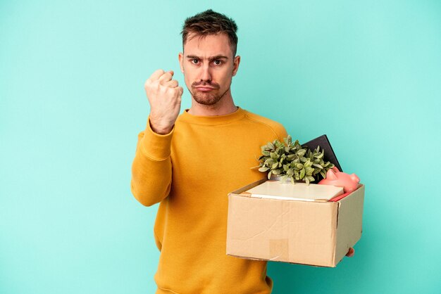 Young caucasian man making a move isolated on blue background showing fist to camera, aggressive facial expression.