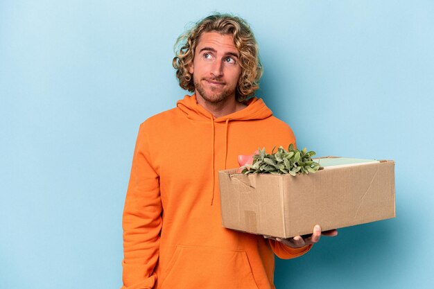 Young caucasian man making a move while picking up a box full of things isolated on blue background dreaming of achieving goals and purposes