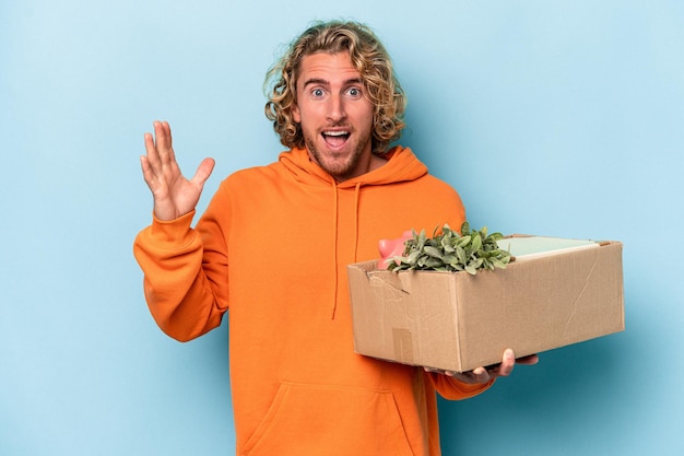 Young caucasian man making a move while picking up a box full of things isolated on blue background receiving a pleasant surprise, excited and raising hands.