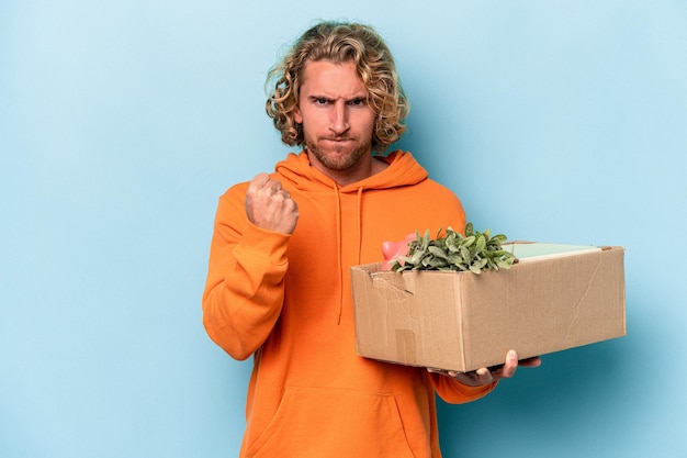 Young caucasian man making a move while picking up a box full of things isolated on blue background showing fist to camera, aggressive facial expression.