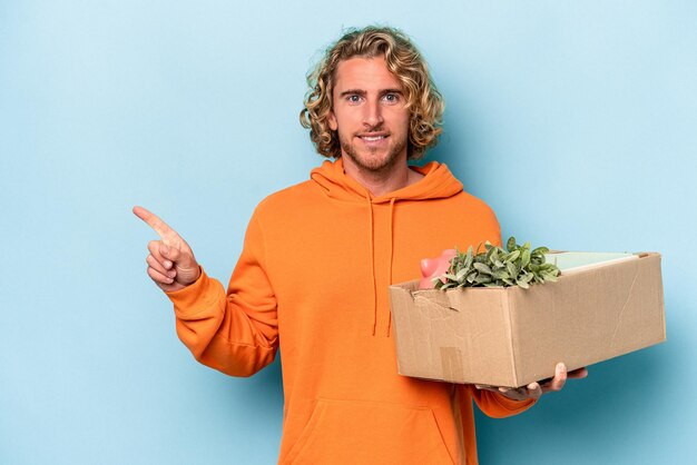 Young caucasian man making a move while picking up a box full of things isolated on blue background smiling and pointing aside, showing something at blank space.