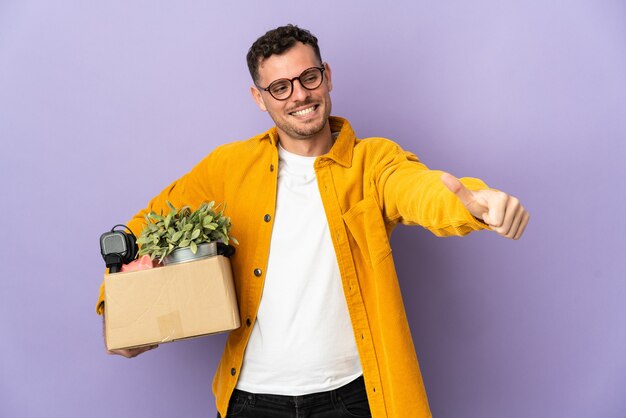 Young caucasian man making a move while picking up a box full of things isolated on purple giving a thumbs up gesture