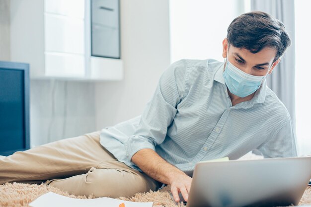 ズボンとシャツのベッドに横たわって、彼のラップトップのキーボードで入力している医療マスクの若い白人男性