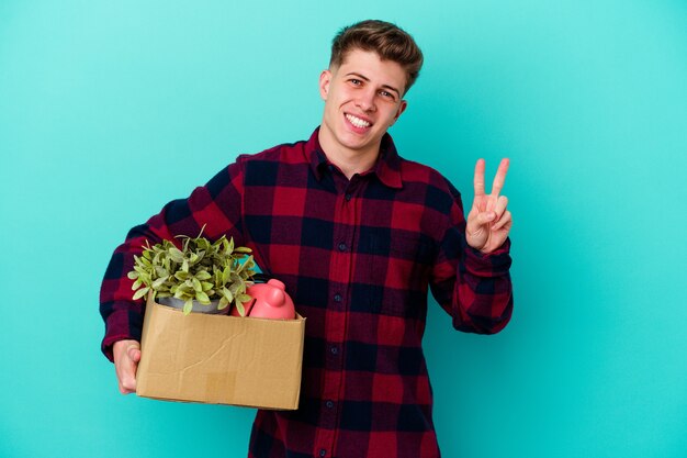 Young caucasian man moving holding a box isolated on blue background joyful and carefree showing a peace symbol with fingers.