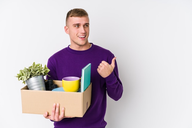 Young caucasian man moving to a new home points with thumb finger away, laughing and carefree.