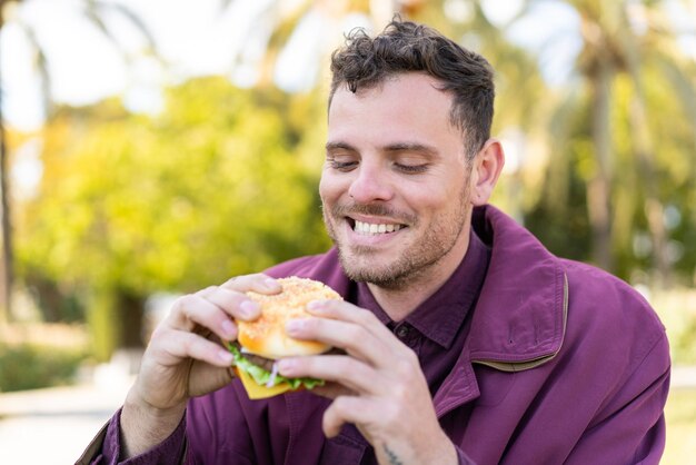 Photo young caucasian man at outdoors holding a burger with happy expression