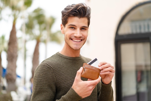 Young caucasian man at outdoors holding wallet and credit card with happy expression